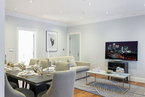 A modern living room with a sofa, dining table set, and a flat-screen TV displaying a cityscape.