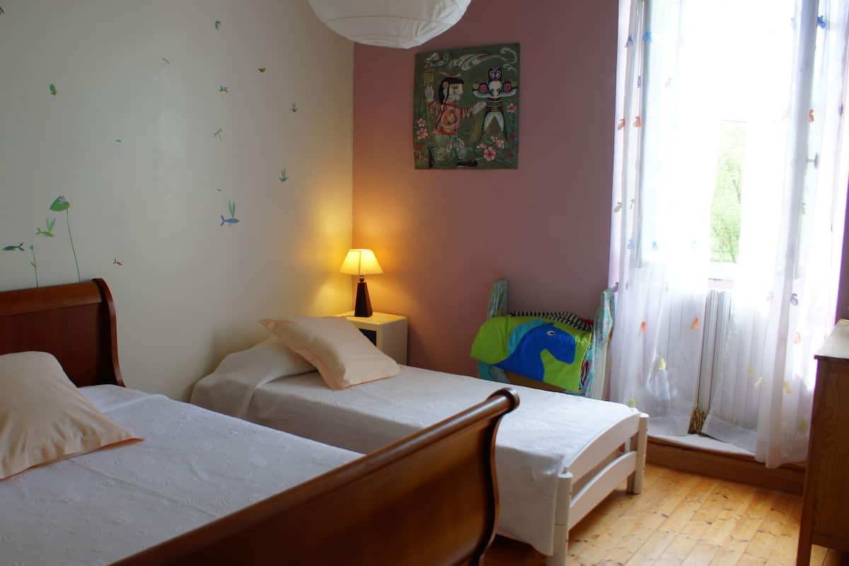 Una habitación con dos camas, una mesita de noche con lámpara, un mural en la pared y una ventana con cortinas.