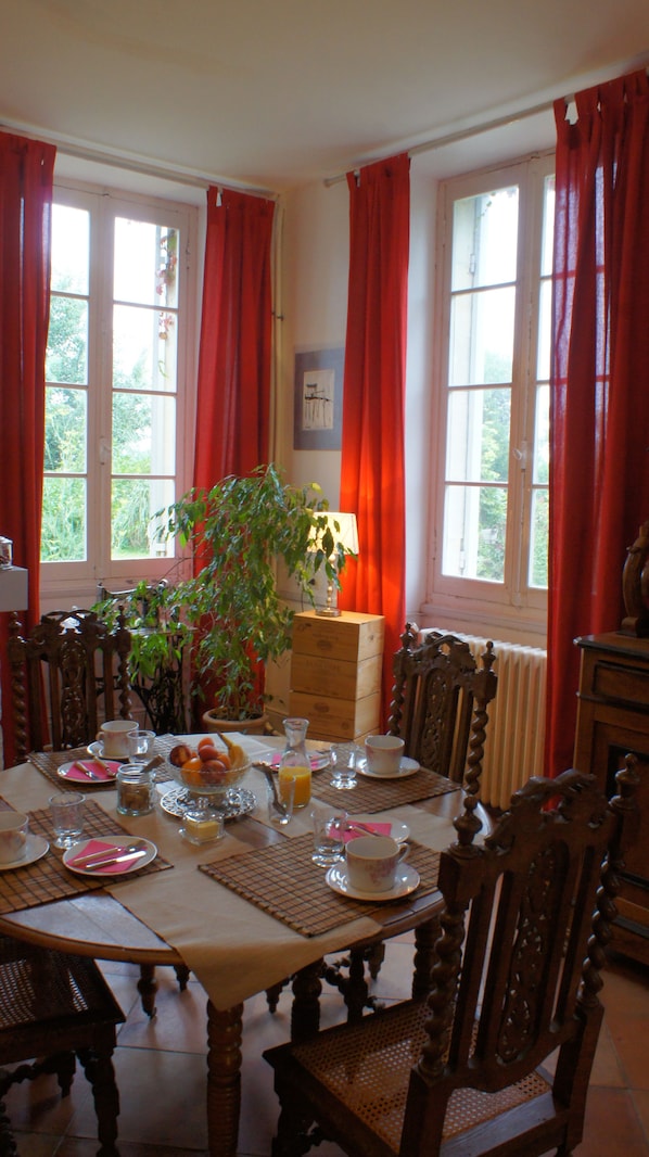 Un comedor con una mesa de madera dispuesta para el desayuno, cortinas rojas y vistas al exterior con vegetación.
