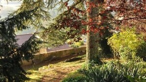 A pathway leading through a garden with a variety of trees and shrubs, a stone wall in the background, and a building with a tiled roof. - Les Jardins de L'hacienda (Tarare)
