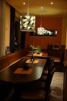 Un comedor con una larga mesa, múltiples sillas, plantas en macetas e iluminación colgante.