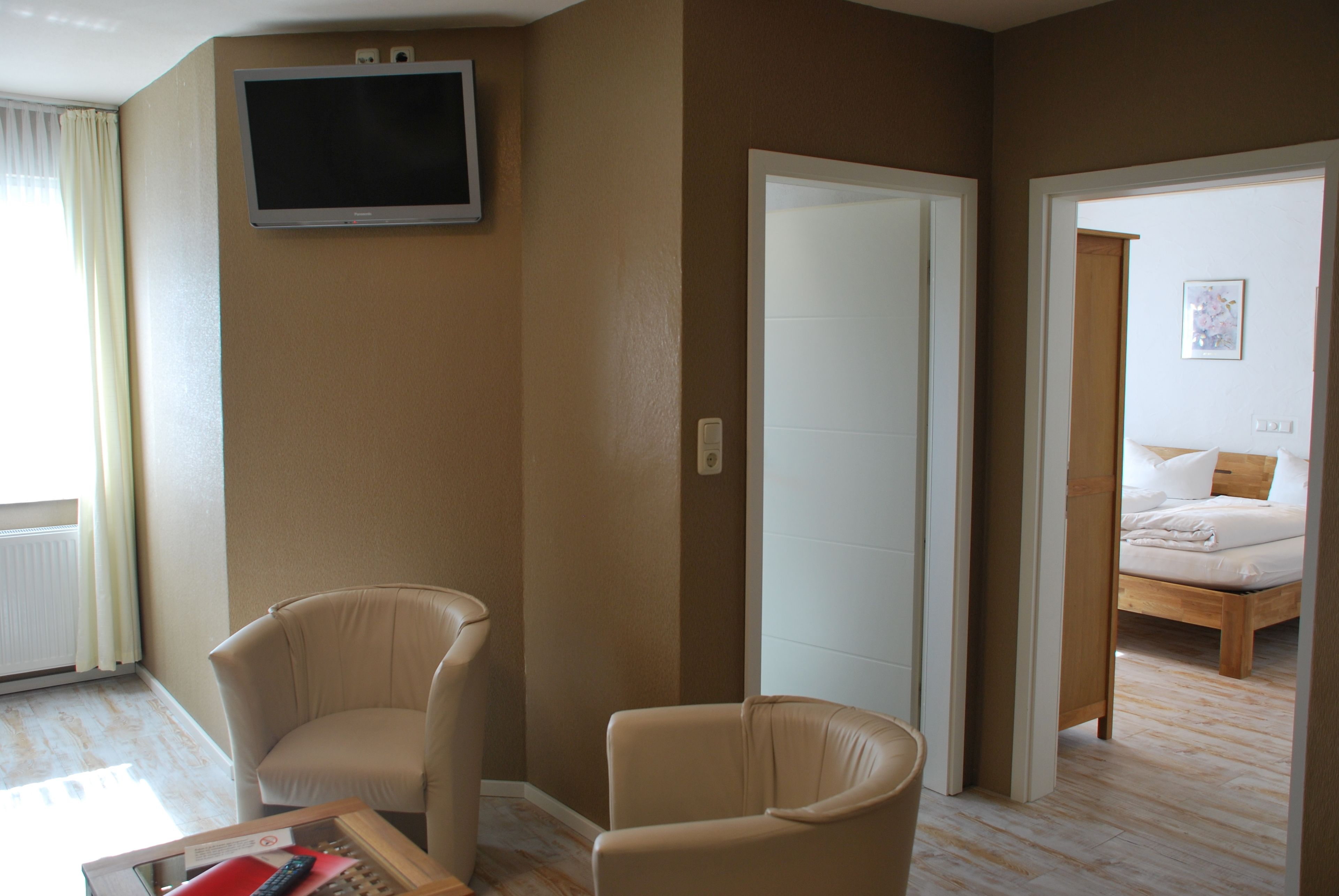 Habitación de hotel con televisor montado en la pared, dos sillas, cama con estructura de madera y puerta que da a otra habitación.