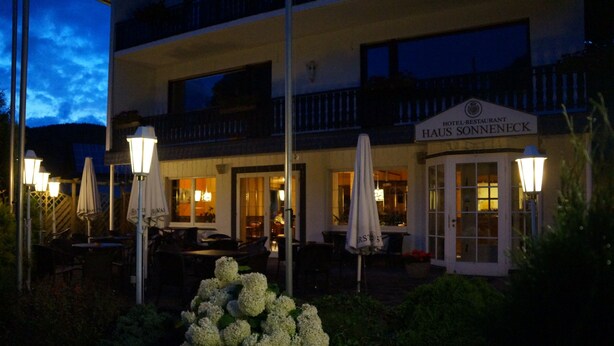 Un hotel con mesas al aire libre y sombrillas, un cartel que dice "HAUS SONNEBECK" y una entrada bien iluminada.