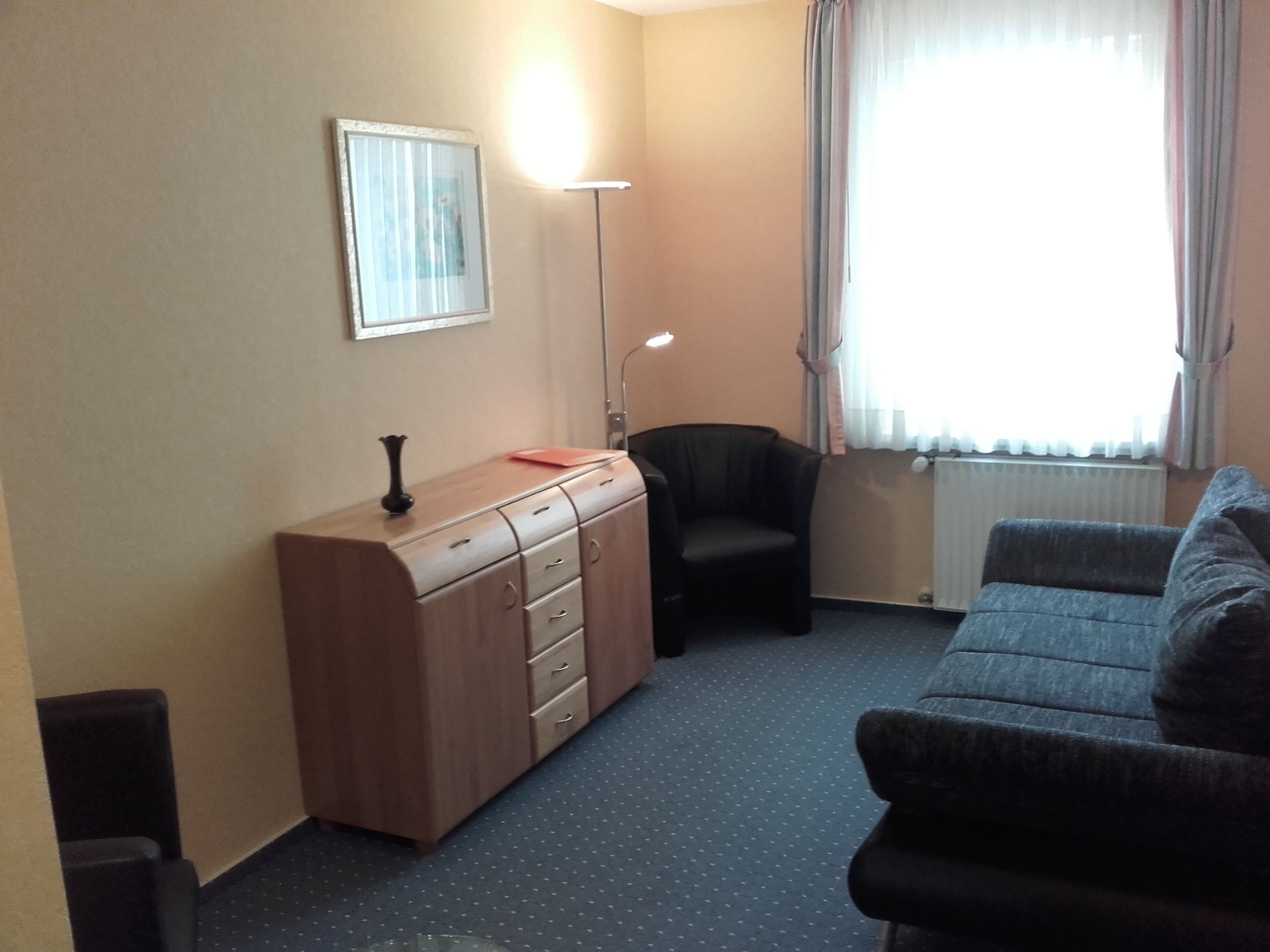 A small, well-lit room with a sofa, a desk with drawers, a chair, a lamp, and a window with curtains.
