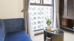 A modern living room with a blue sofa, a glass coffee table, and a view of a cityscape through large windows. - Blubiz Hotel My Dinh Song Da (Hanoi)