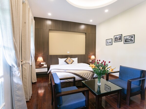 Habitación de hotel con una cama, mesitas de noche, una mesa de centro de vidrio, dos sillas, un jarrón con flores y cuadros en la pared.