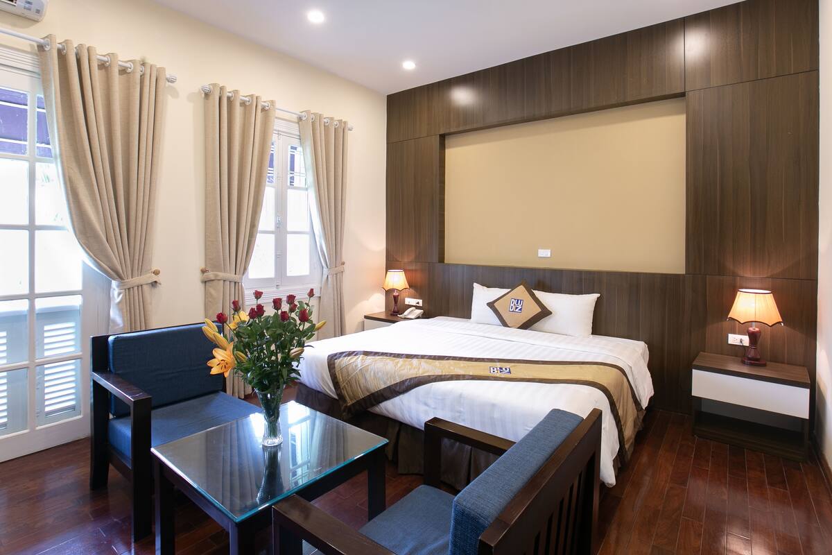 A hotel room with a bed, two bedside tables, a small table with a vase of flowers, a sofa, and a chair.