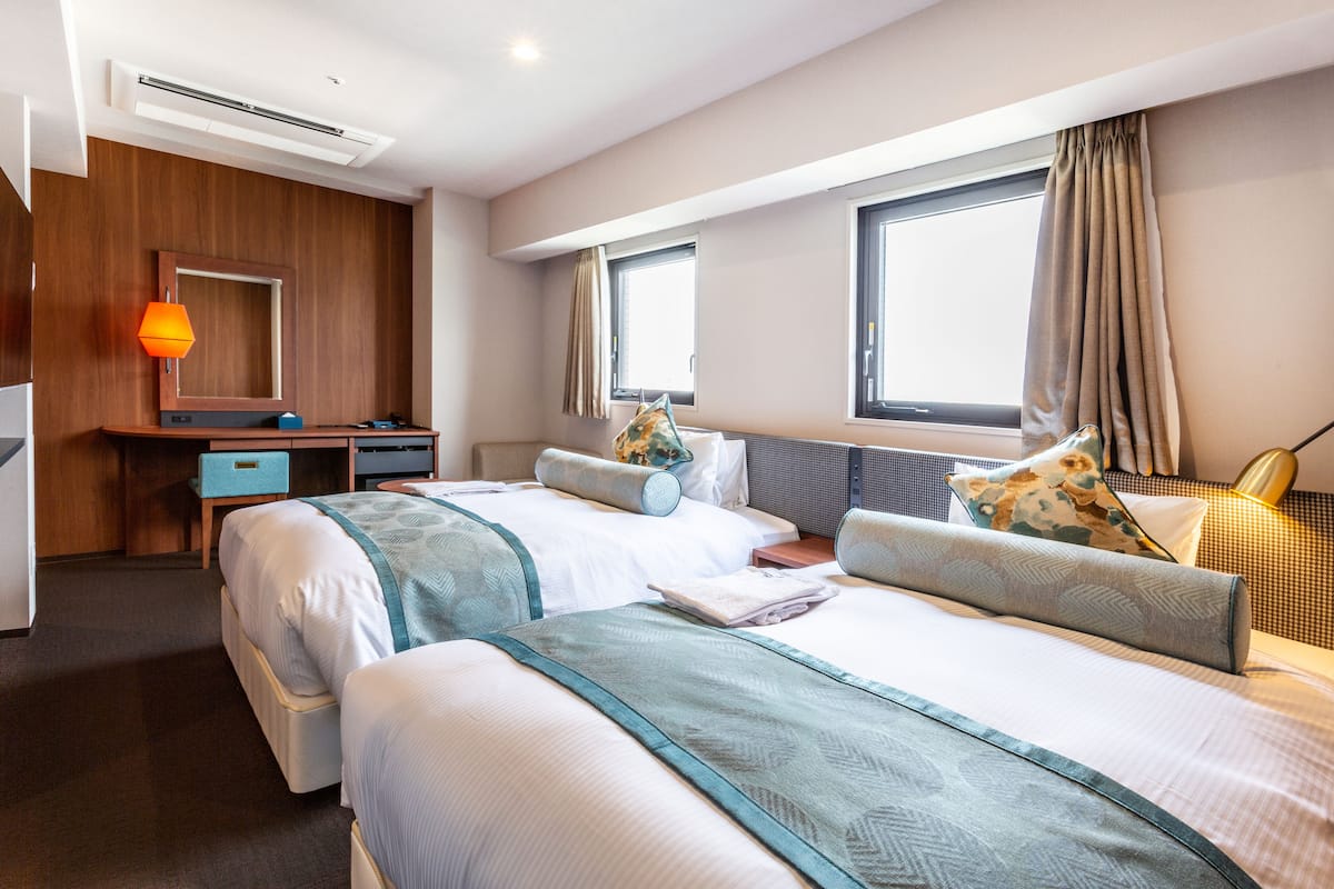 A modern hotel room with two beds, a desk, a chair, a lamp, and large windows.
