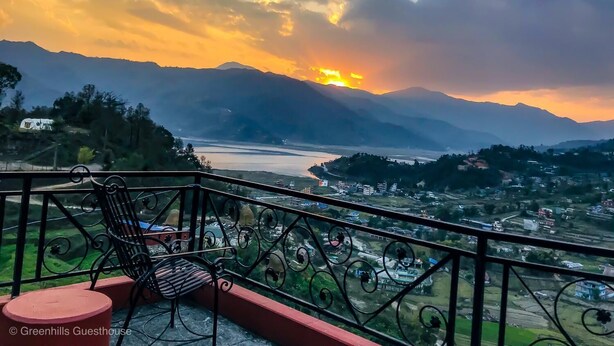 A balcony with a chair and a small table, overlooking a valley with mountains and a river.