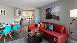 A modern living room with a red sofa, a glass coffee table, and a dining area with blue chairs. - Green Diamond by Creatick Apartments (Brighton)