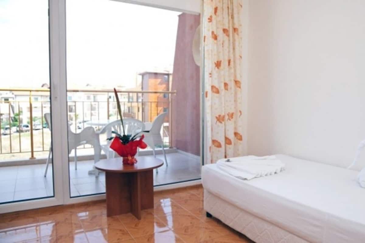 A hotel room with a bed, a small table with a plant, and a view of the outside balcony.