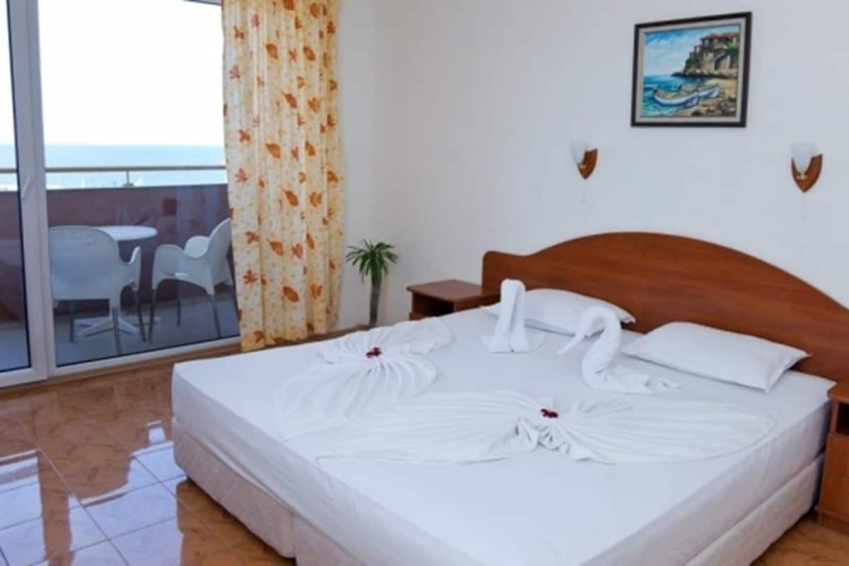 A hotel room with a bed, a nightstand, a small table, a chair, and a view of the sea through a glass door.