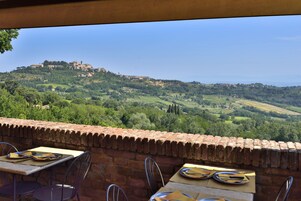 Un área de comedor al aire libre con mesas y sillas, con vistas a un paisaje verde exuberante y una colina con edificios.
