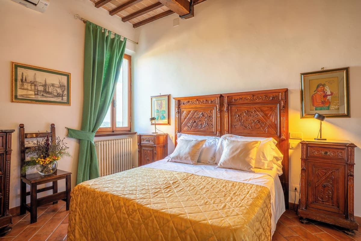 Een slaapkamer met een groot bed, een houten hoofdbord, een nachtkastje, een lamp en een raam met groene gordijnen.