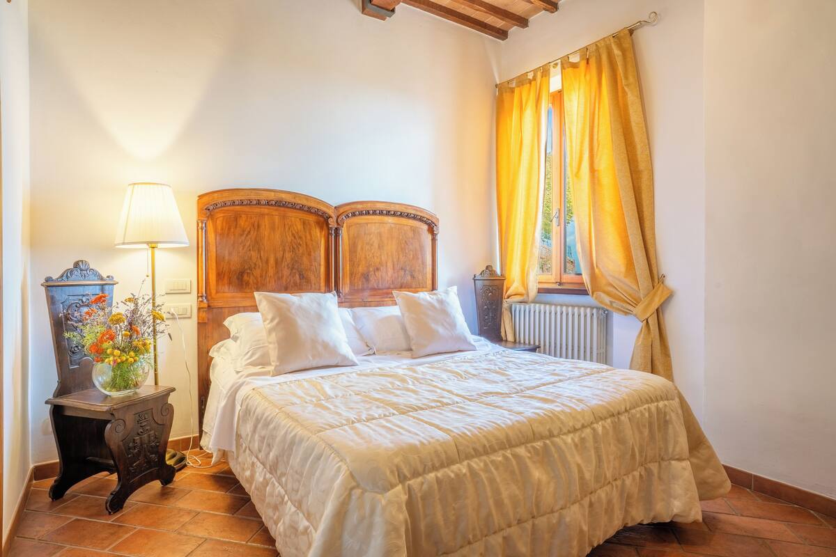 Een slaapkamer met een houten hoofdbord, een bed met witte beddengoed, een nachtkastje met een lamp en een raam met gele gordijnen.