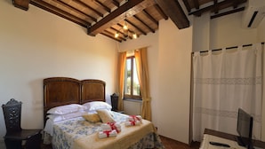 A bedroom with a wooden bed, a nightstand, a desk, a window with curtains, a radiator, and a television on a table. - La Corte Medicea (Montepulciano)