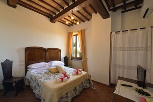 Un dormitorio con cama de madera, mesita de noche, escritorio, ventana con cortinas, radiador y televisor sobre una mesa.