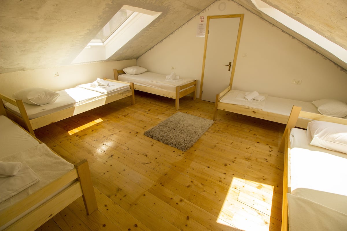 Una habitación pequeña tipo ático con tres literas de madera dispuestas en forma de U, una puerta y un tragaluz.