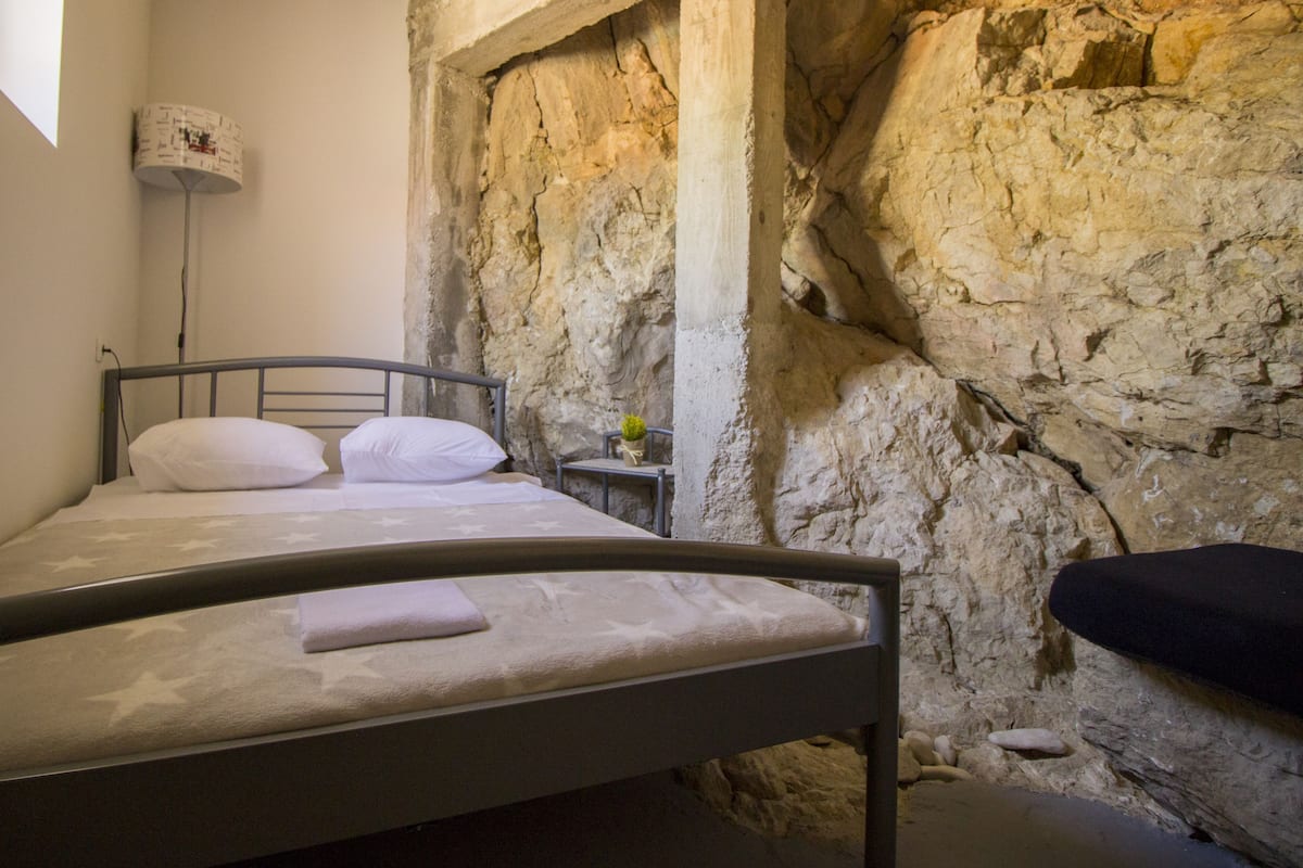 Una cama en una habitación con pared de piedra, una mesita de noche con lámpara y una planta pequeña.