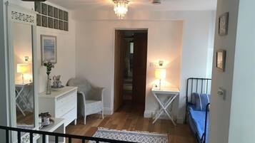 A room with a wooden floor, a white dresser, a chair, a small table with a lamp, and a blue sofa.