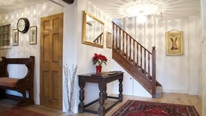 Um interior tradicional com uma escadaria de madeira, um banco de madeira, uma mesa com um vaso de flores e um relógio na parede.