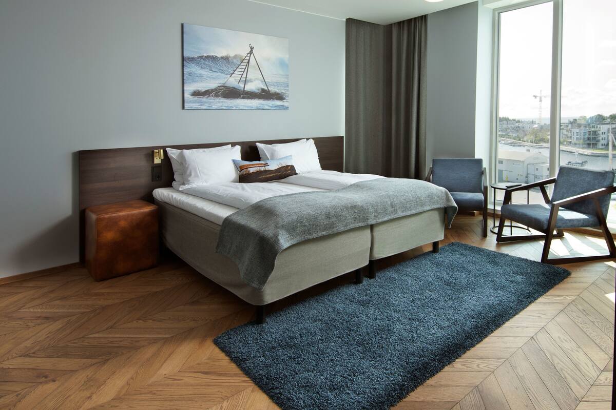 A modern hotel room with a large bed, a view of the city through the window, and a painting of a ship on the wall.