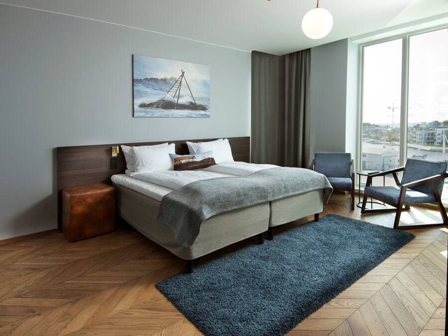 A modern hotel room with a large bed, a view of the city through the window, and a painting of a ship on the wall.