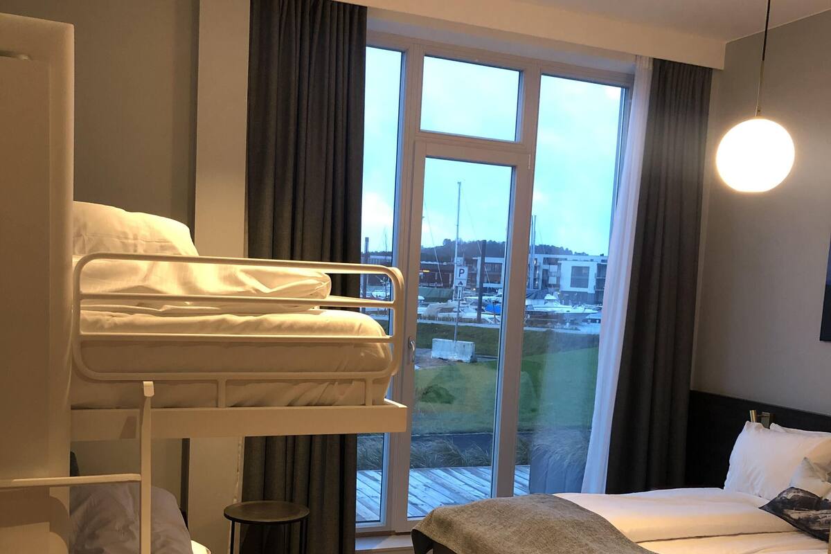 A hotel room with a bunk bed, a single bed, a small table, and a view of the outside.