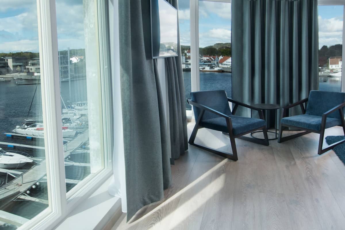 A hotel room with a large window overlooking a marina, a bed with a blue throw, two armchairs, and a small table.