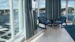 A hotel room with a large window overlooking a marina, a bed with a blue throw, two armchairs, and a small table.