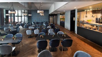 A spacious dining area with multiple tables and chairs, a modern kitchen visible through a glass partition, and pendant lighting.