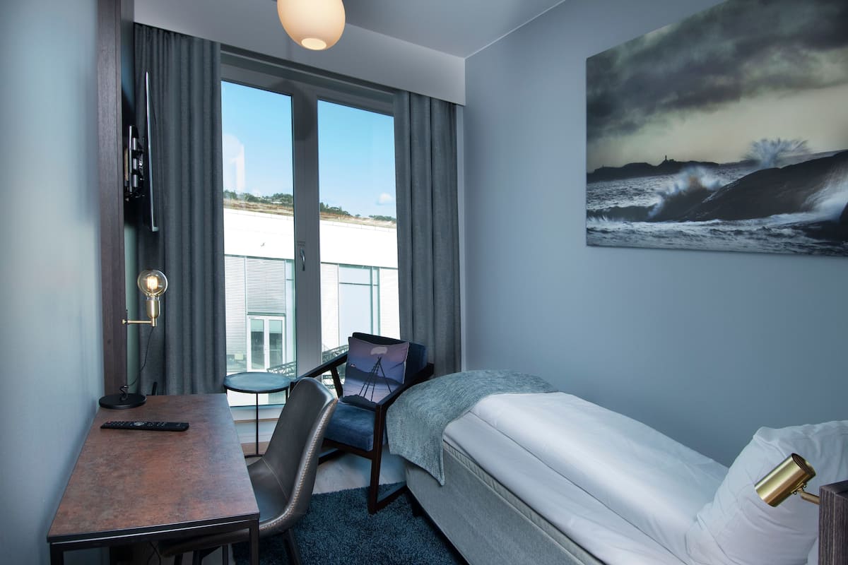 A hotel room with a bed, a desk with a chair, a small table, a remote control, and a painting of a stormy sea on the wall.
