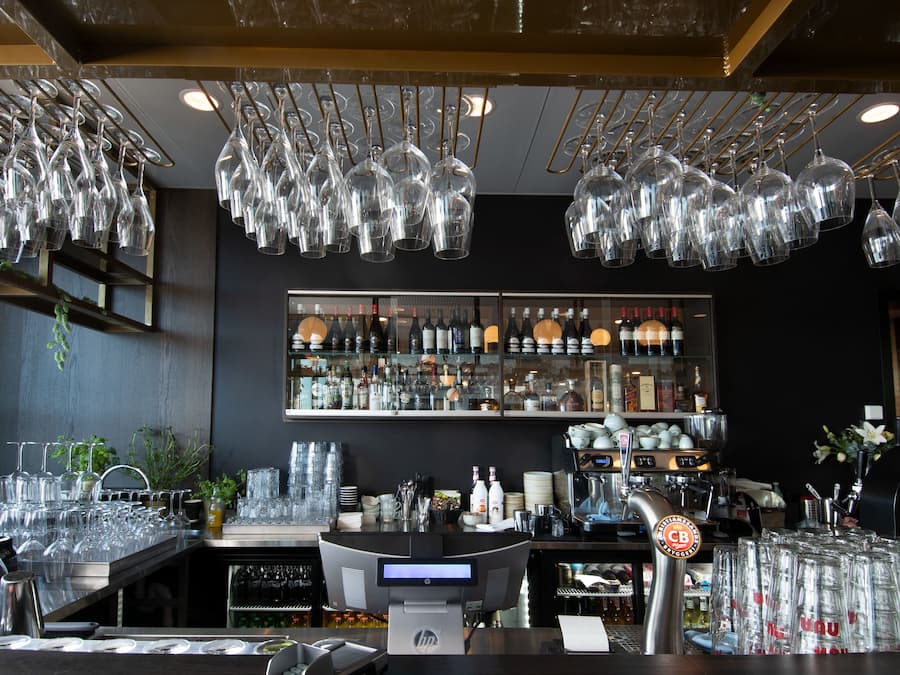 Zona de bar con varios vasos de vino colgando del techo, una barra con una caja registradora y estantes llenos de botellas y copas.
