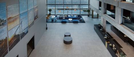 A spacious lobby with a high ceiling, large windows overlooking a body of water, and a seating area with round tables and chairs.