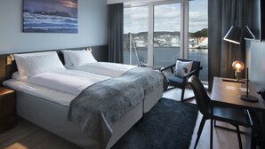A modern hotel room with a large bed, a desk with a lamp, a chair, and a view of the water and buildings. - Mandal Hotel (Lindesnes)