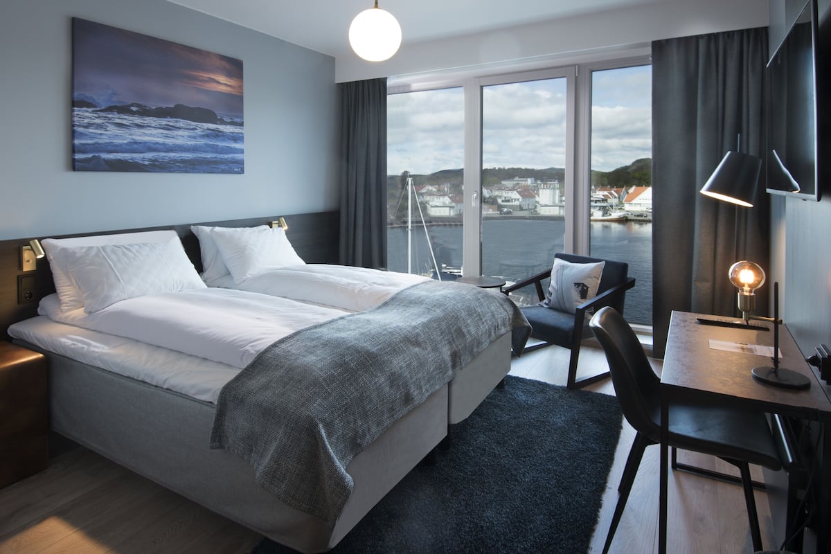 A modern hotel room with a large bed, a desk with a lamp, a chair, and a view of the water and buildings.