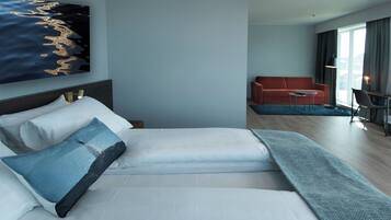 Ett modernt hotellrum med en stor säng, en soffa, ett litet bord och utsikt över staden genom stora fönster.
