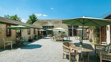 An outdoor patio area with tables and chairs, surrounded by brick buildings and thatched-roof structures.