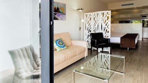 Una sala moderna con un sofá, una mesa de centro de vidrio y una silla negra. Se observa una cama con cabecera marrón al fondo.