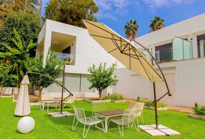Un espace extérieur moderne, aménagé avec des chaises et des tables en métal blanc, entouré de gazon synthétique vert et de palmiers.