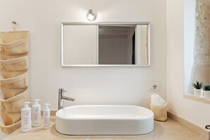 Une salle de bain avec une grande baignoire blanche, un miroir au-dessus et un porte-serviettes avec une serviette beige. Trois bouteilles de shampoing sont posées sur le sol.