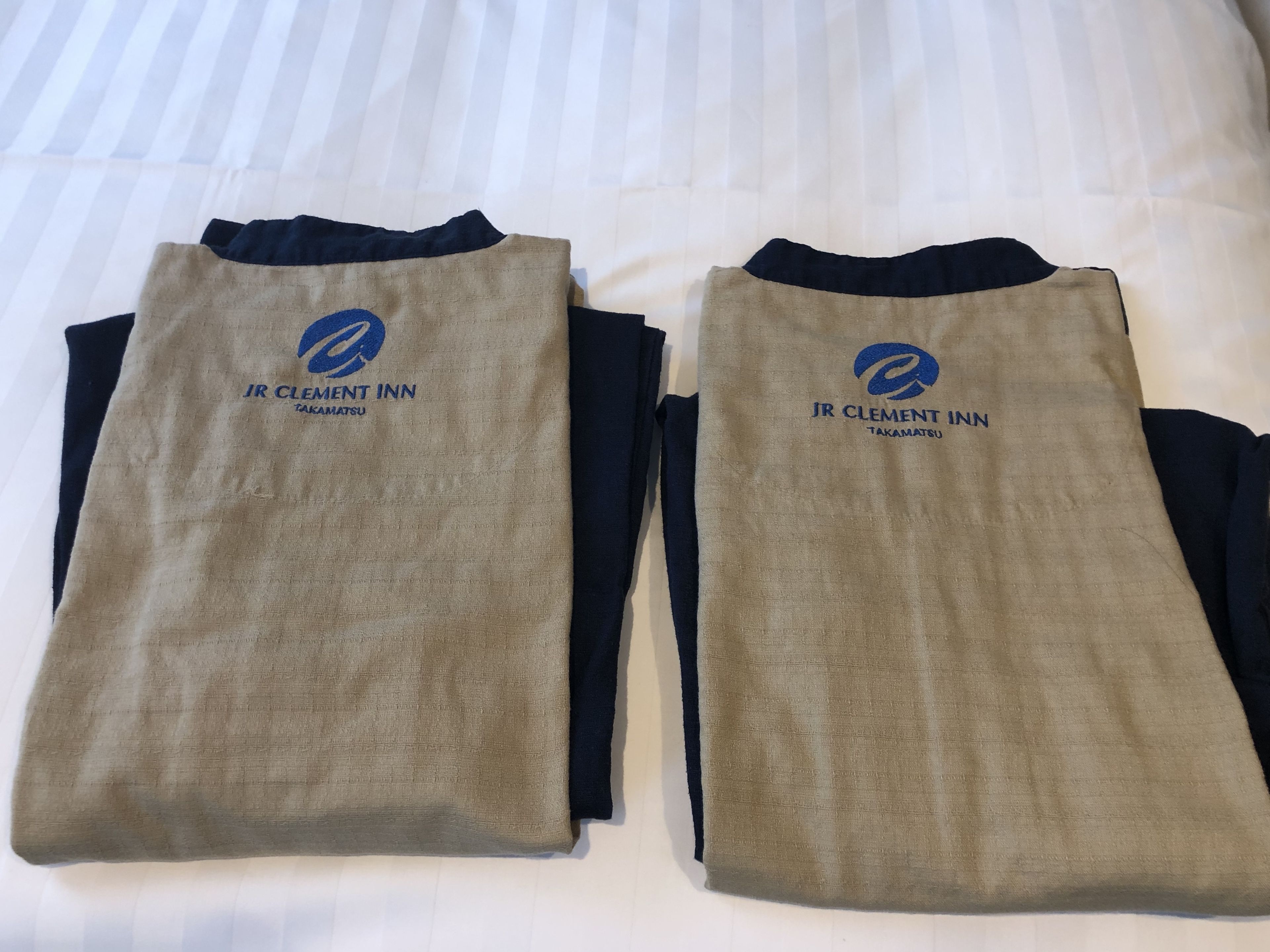 Dos camisas de manga larga con el logotipo y el nombre de JR Clement Inn Takamatsu, extendidas sobre una cama.
