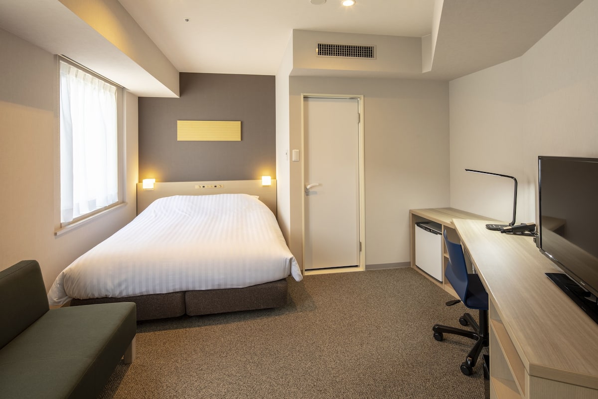 Une chambre d’hôtel avec un lit, un bureau, une chaise, une télévision et une fenêtre avec des rideaux.
