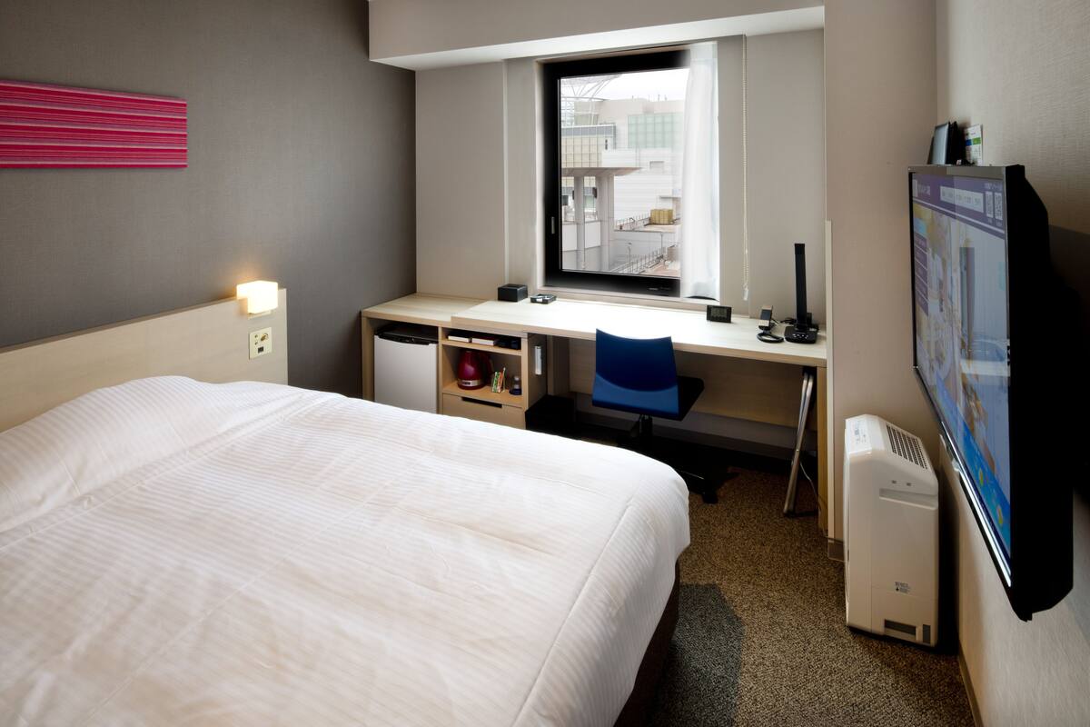 Une chambre d’hôtel avec un lit, un bureau avec un ordinateur, une chaise et une fenêtre.