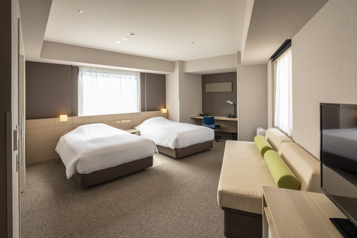 Une chambre d’hôtel avec deux lits, un canapé, une télévision, un bureau et une fenêtre avec des rideaux.