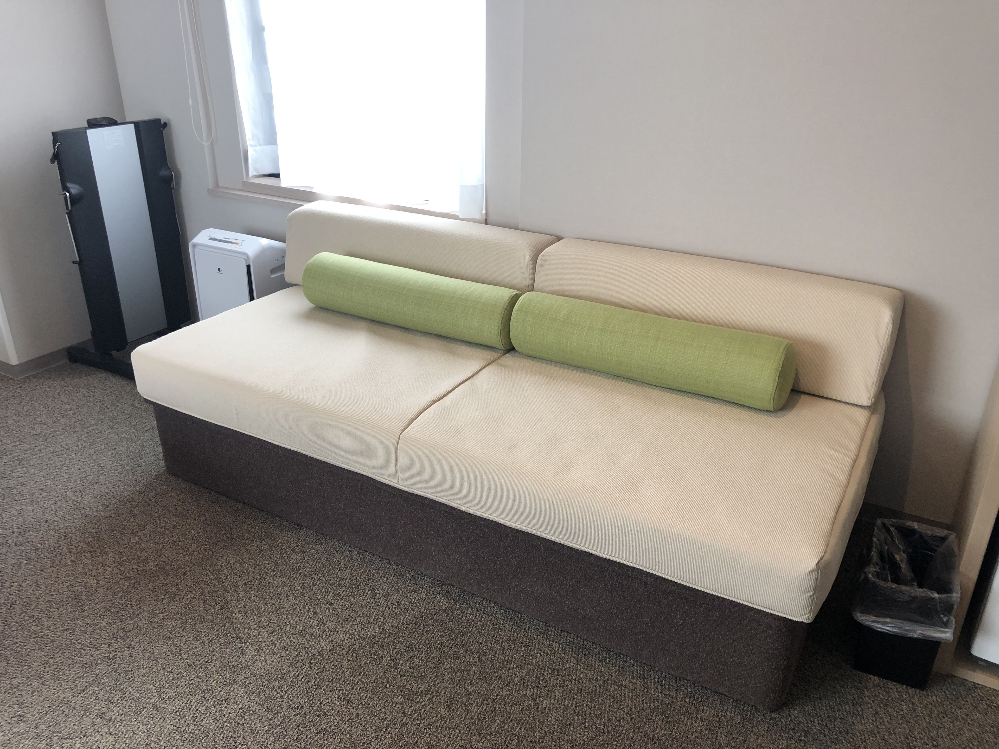 A beige sofa with a green pillow, a small trash bin, and a window with white curtains.