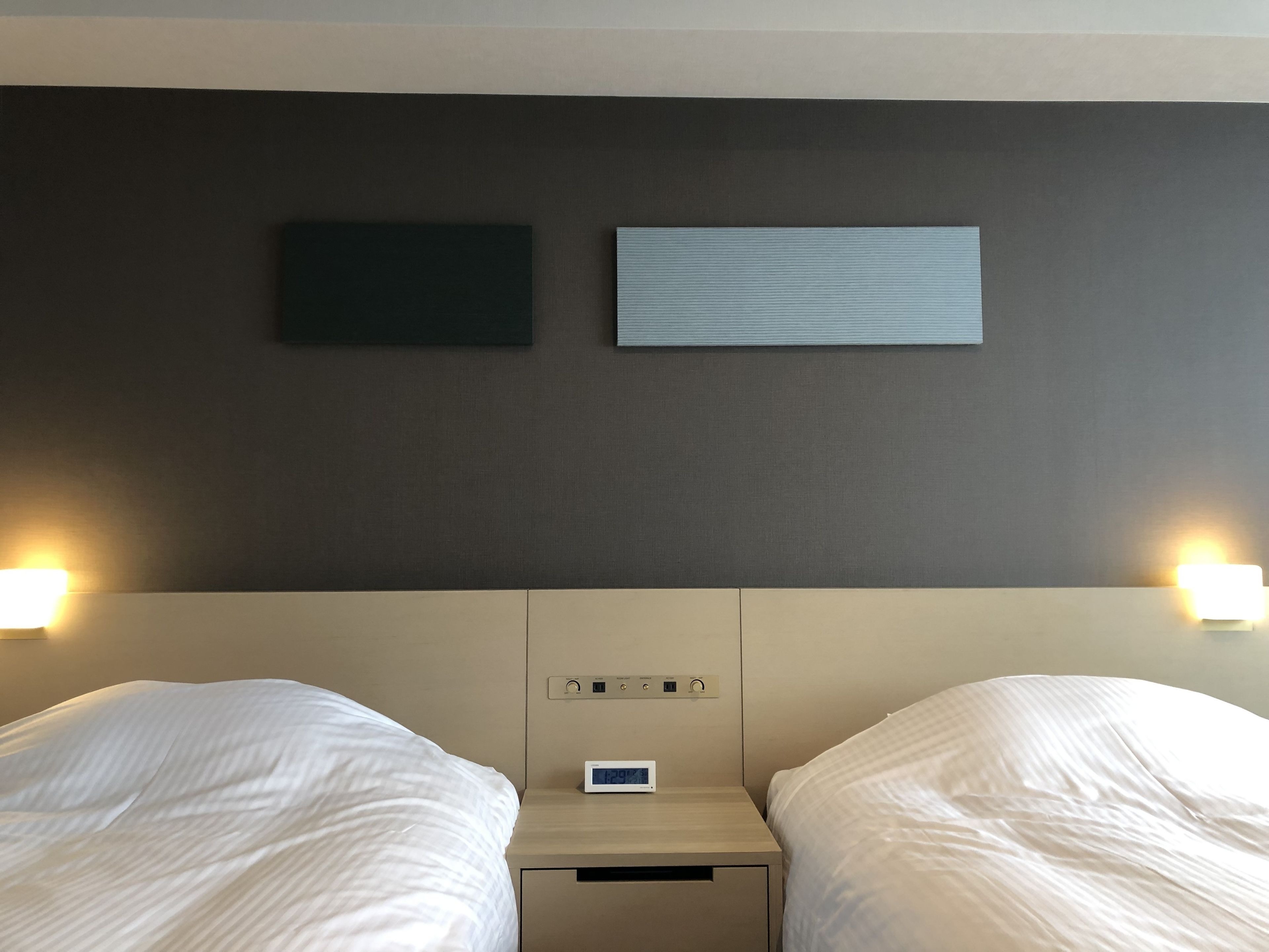 A hotel room with two beds, a nightstand with a clock, and wall-mounted lights.