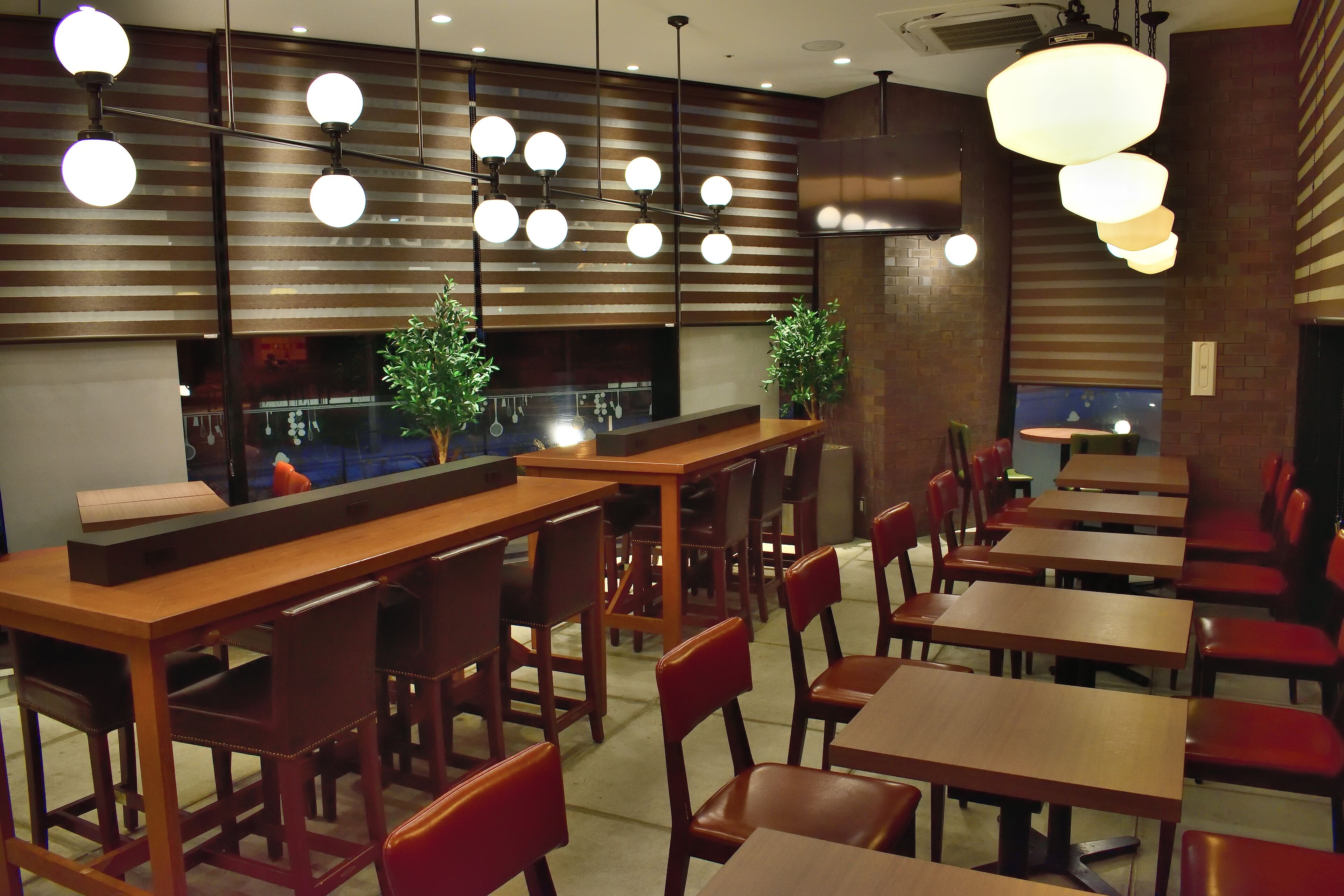 A restaurant interior with multiple wooden tables and red chairs, hanging spherical light fixtures, and a brick wall backdrop.