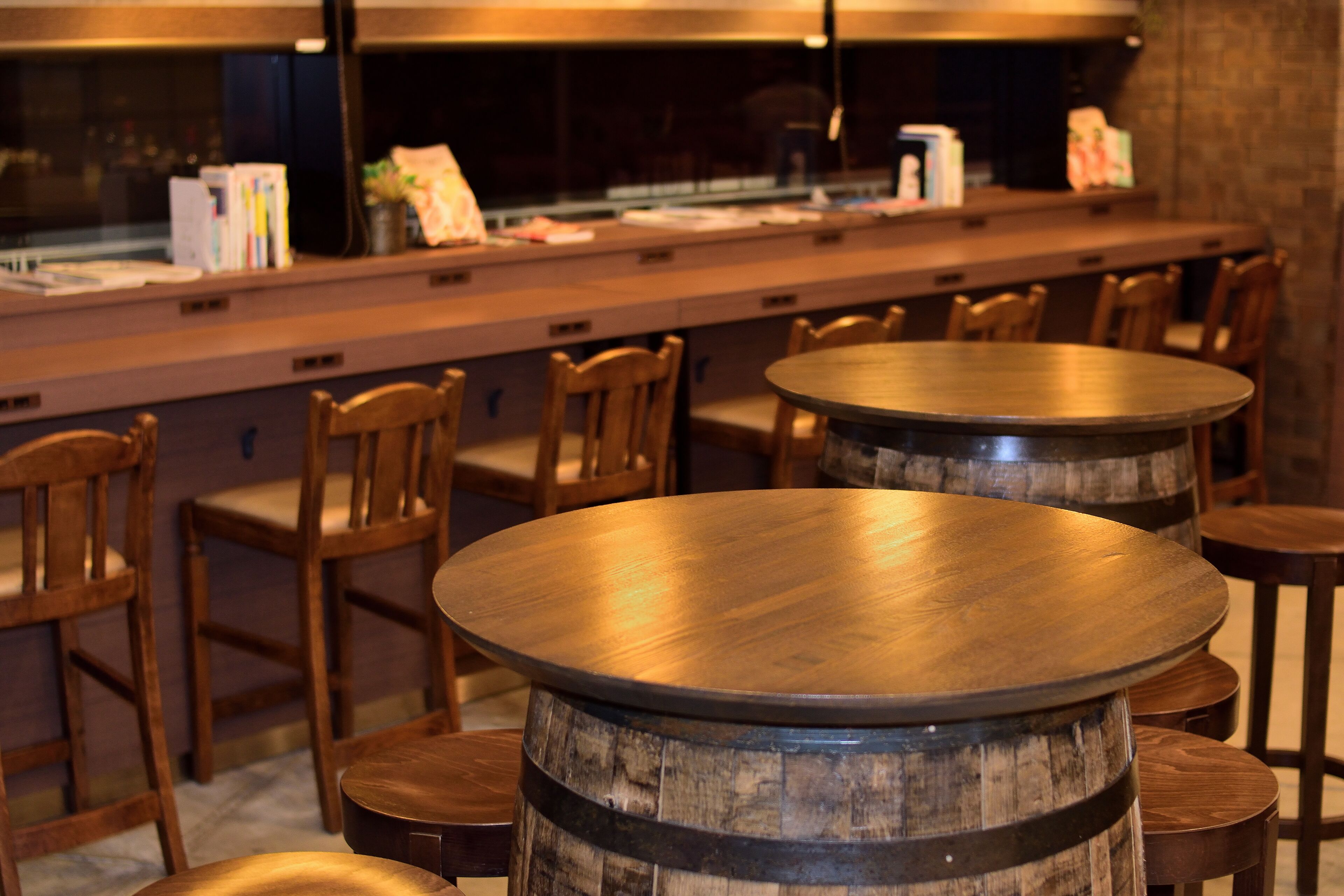 Una zona de bar con mesas y sillas de madera, una barra con libros y papeles, y una ventana al fondo.