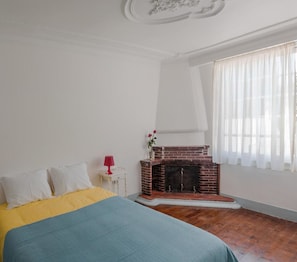 Une chambre à coucher avec un lit, une cheminée, une fenêtre ornée de rideaux légers et un plafond blanc agrémenté de moulures décoratives.